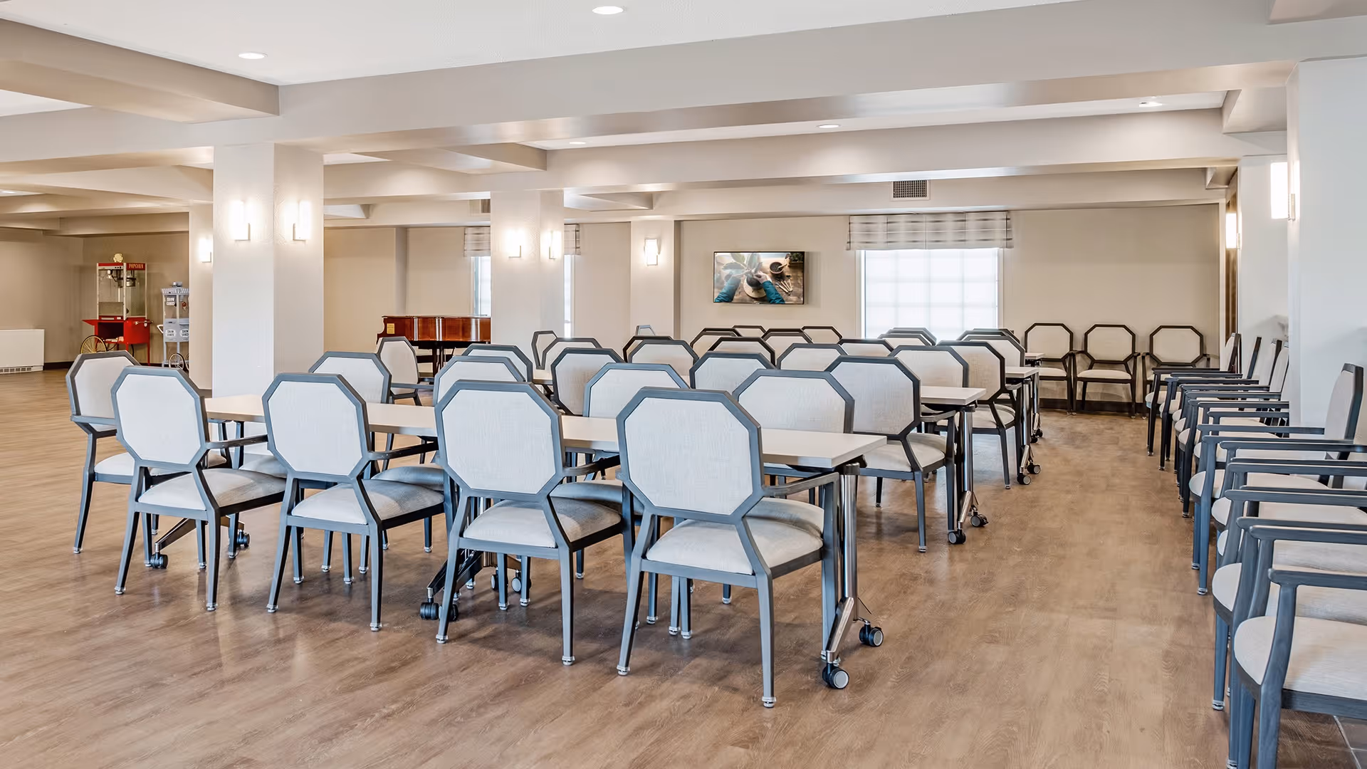 A spacious dining room with multiple tables and chairs arranged in rows on a wooden floor. The room has white walls, ceiling lights, and a large window with a blind. There is a popcorn machine and a piano in the background.