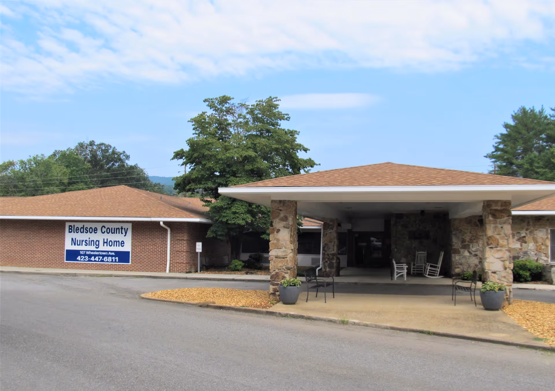 Exterior view of Bledsoe County Nursing Home showing a single-story brick building with a covered entrance supported by stone pillars. There are chairs and potted plants under the entrance canopy, and a sign on the building displays the facility's name, address, and phone number.