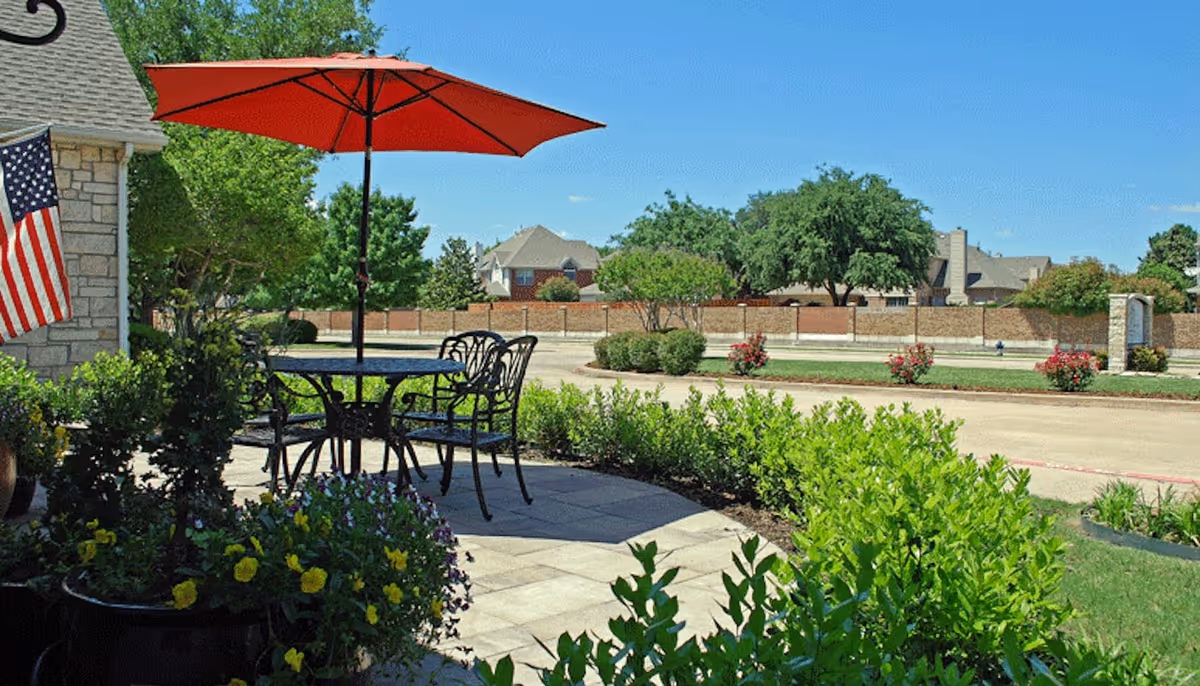 Patio with a table, chairs and a red umbrella overlooking landscaped greenery and a street with neighboring houses.