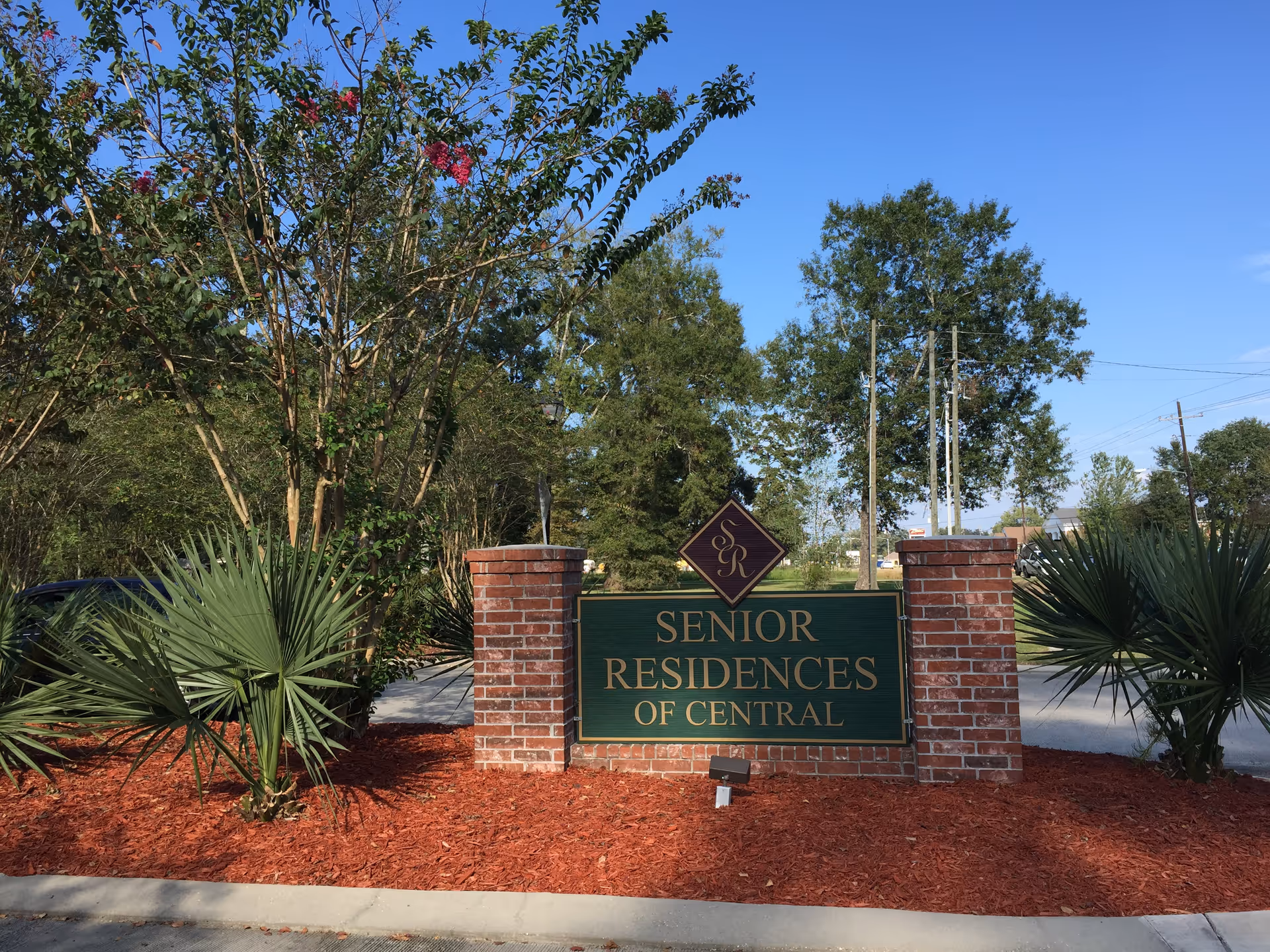 Outdoor view of a sign for Senior Residences of Central, mounted on a brick base with landscaping including small palm plants and trees under a clear blue sky.
