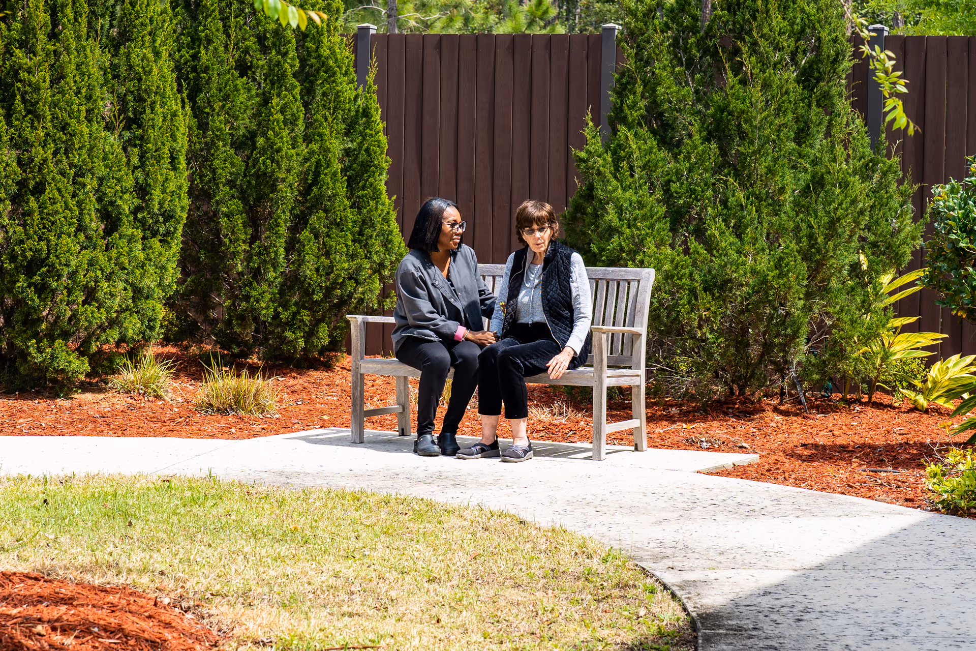 Two women sitting on a wooden bench in a garden area with green shrubs and a brown wooden fence in the background. One woman is wearing a gray jacket and black pants, and the other is wearing a black vest over a gray shirt and black pants. They appear to be engaged in conversation.