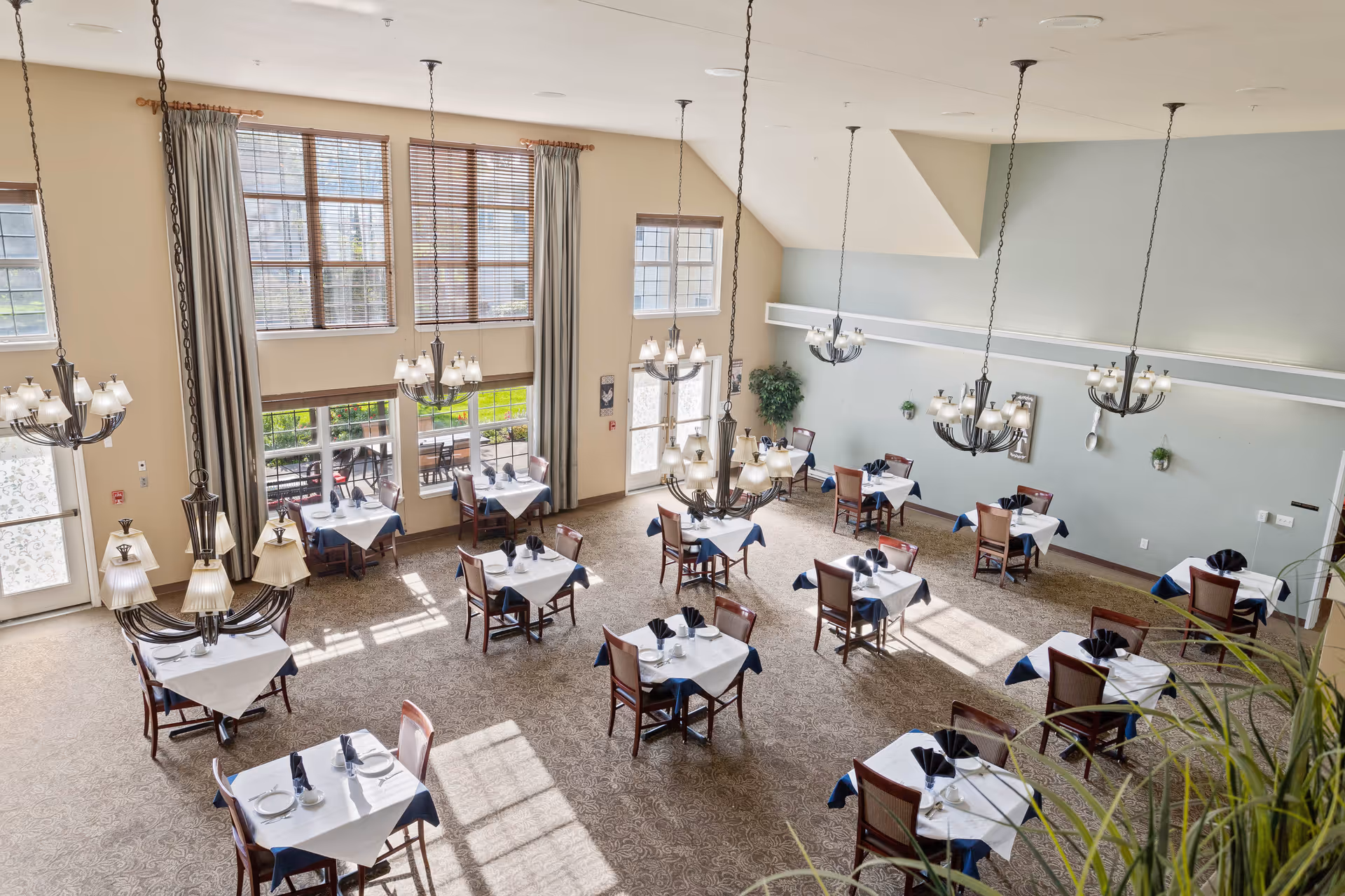 A bright and spacious dining room with multiple tables set with white tablecloths and dark napkins. The room features large windows with curtains, several hanging chandeliers, and carpeted flooring. Chairs are neatly arranged around each table, and there are some plants and wall decorations on the light-colored walls.