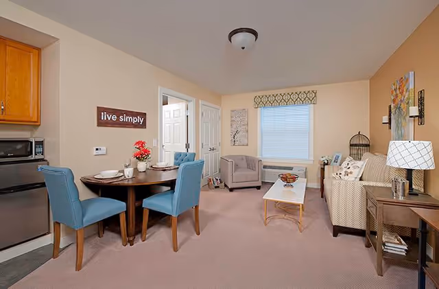 A cozy living and dining area in a senior living facility with beige walls and carpet. The dining area has a round wooden table set with plates, cups, and a small flower arrangement, surrounded by four blue upholstered chairs. The living area features a beige patterned sofa, a cushioned armchair, a white coffee table with a decorative bowl, and a side table with a lamp. A window with a patterned valance lets in natural light. Wall decor includes a 'live simply' sign and framed artwork.