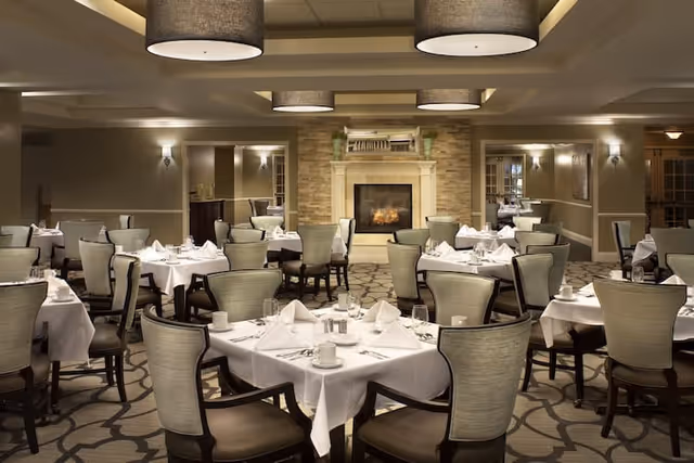 Dining room with neatly set tables and upholstered chairs centered around a fireplace under large pendant lights.