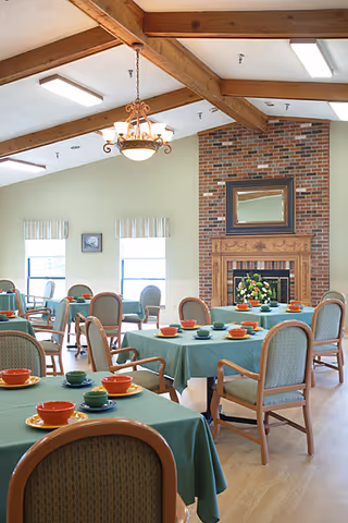 Sunlit dining room with multiple tables covered in green tablecloths set with colorful bowls and cups, exposed wooden beams, and a brick fireplace.