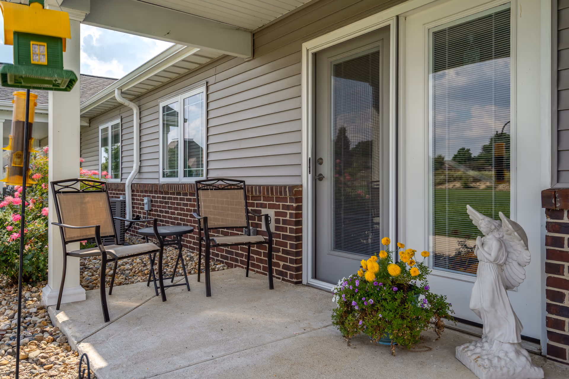Outdoor patio area with two metal chairs and a small round table on a concrete floor. There is a flower pot with yellow and purple flowers next to a white statue of an angel. The patio is adjacent to a building with gray siding and brick accents, featuring a glass door and window with blinds. A bird feeder hangs from a white post on the left side.