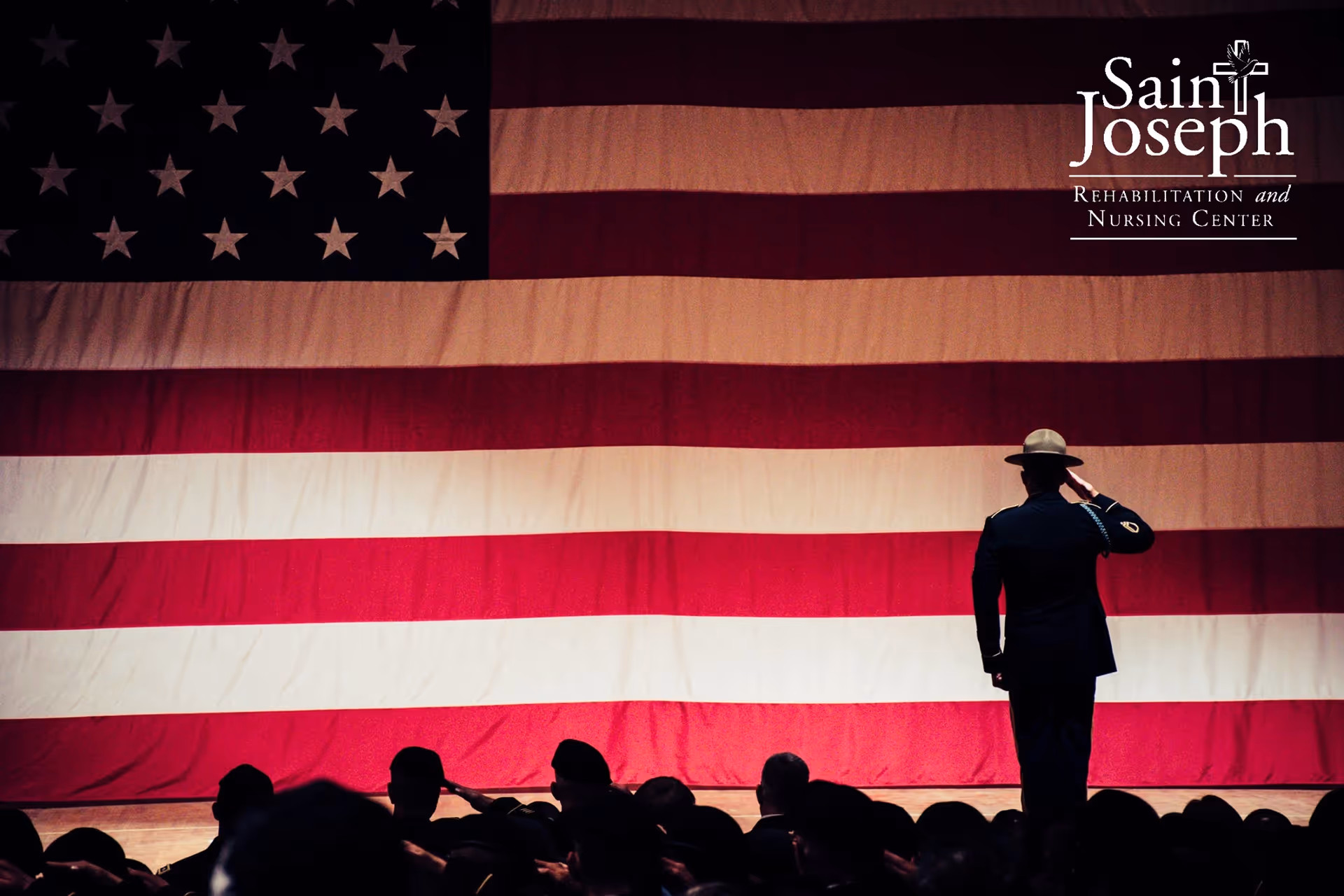 A uniformed person salutes in front of a large American flag with audience silhouettes in the foreground.