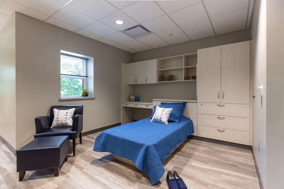 A simple, clean bedroom with a single bed covered in a blue quilt and matching pillows with anchor designs. There is a black armchair with a matching ottoman and an anchor pillow next to a window with blinds. The room has light wood flooring, built-in light wood cabinets and shelves, and a pair of blue slippers on the floor near the bed.
