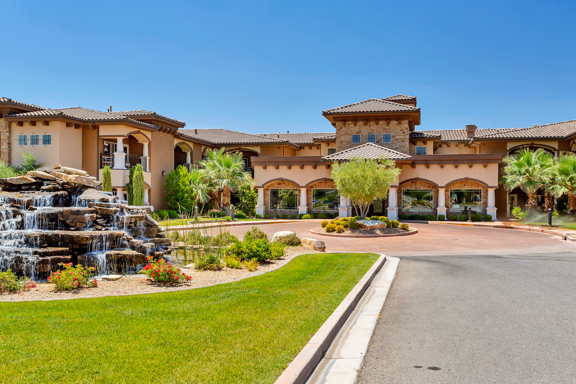 Front exterior view of The Retreat at Sunbrook facility featuring a large building with a tiled roof, arched windows, and a landscaped entrance with palm trees, bushes, and a rock waterfall feature under a clear blue sky.