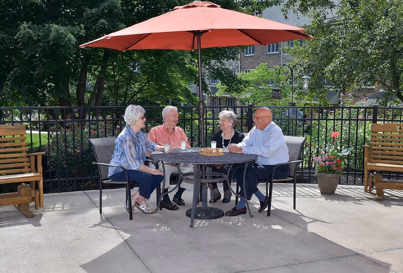 Four elderly people sitting around a round outdoor table under a large red umbrella, enjoying drinks and conversation on a sunny patio with greenery and a black metal fence in the background.