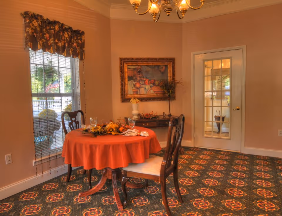 A cozy dining room with a round table covered by an orange tablecloth, set with decorative fall-themed centerpiece and two wine glasses. Two wooden chairs with cushioned seats are placed around the table. A window with floral valance and blinds is on the left, and a glass-paneled door is on the right. The room has patterned carpet and a framed painting on the wall above a small console table with decorative items.