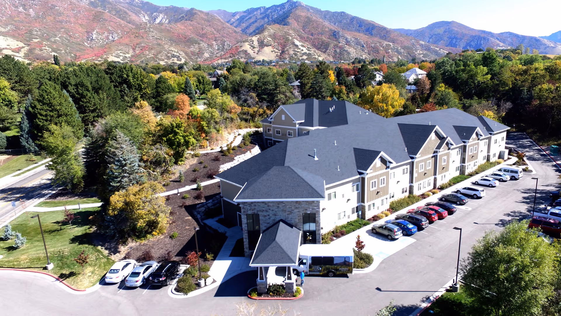 Aerial view of a large assisted living and memory care facility named Hidden Valley, surrounded by trees and mountains in the background. The building has a dark roof and light-colored walls with a parking lot filled with cars on the right side. There are landscaped gardens and pathways around the building.