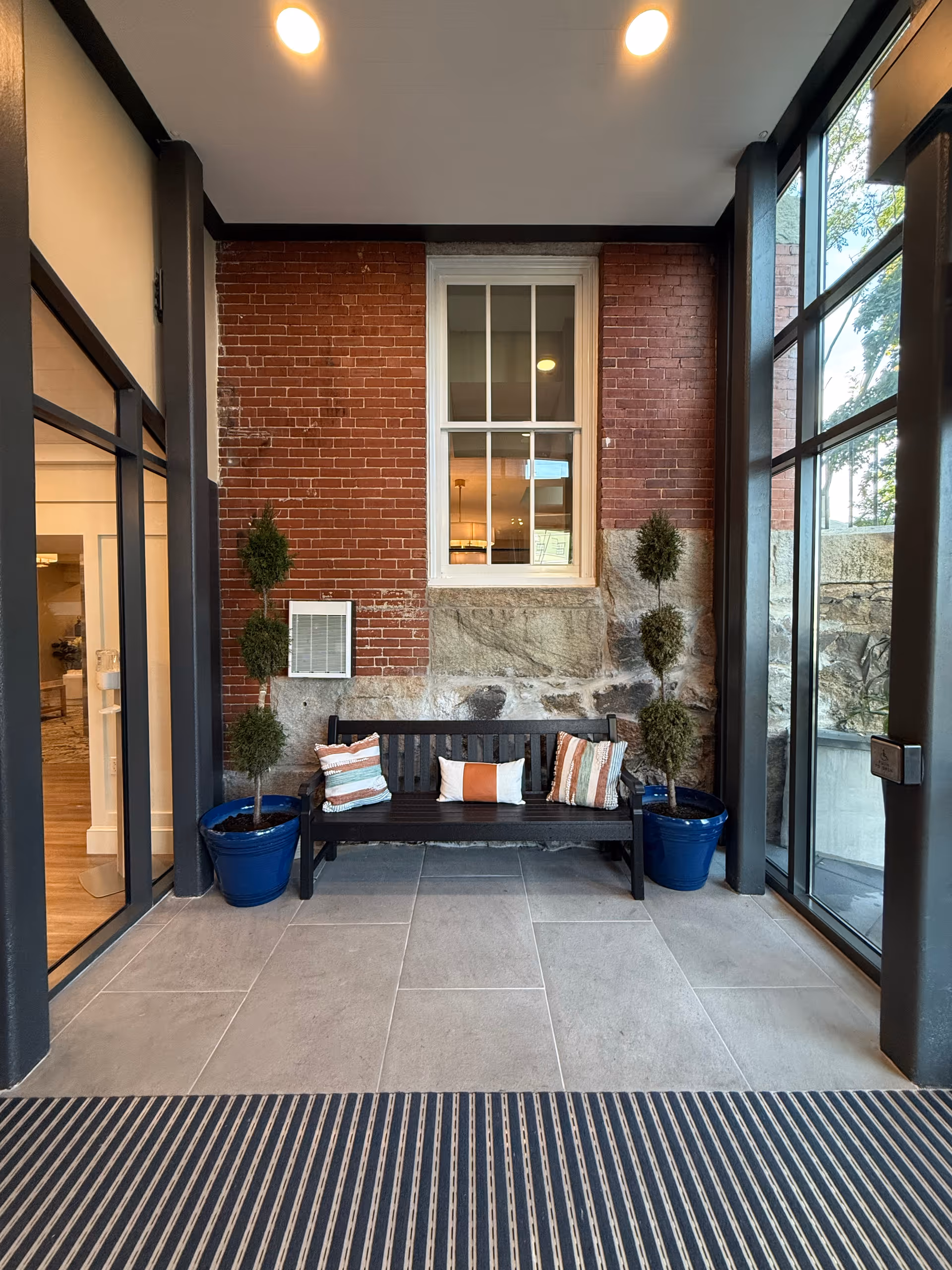 Indoor entryway with a black bench against a brick and stone wall, potted topiary plants, and a window flanked by glass doors.