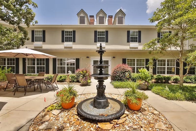 Outdoor courtyard area of a senior living facility with a three-tiered black fountain in the center surrounded by rocks and potted plants. There is a patio with a table, chairs, and an umbrella to the left. The building in the background is two stories with multiple windows and dormer vents on the roof. Trees and shrubs frame the courtyard.