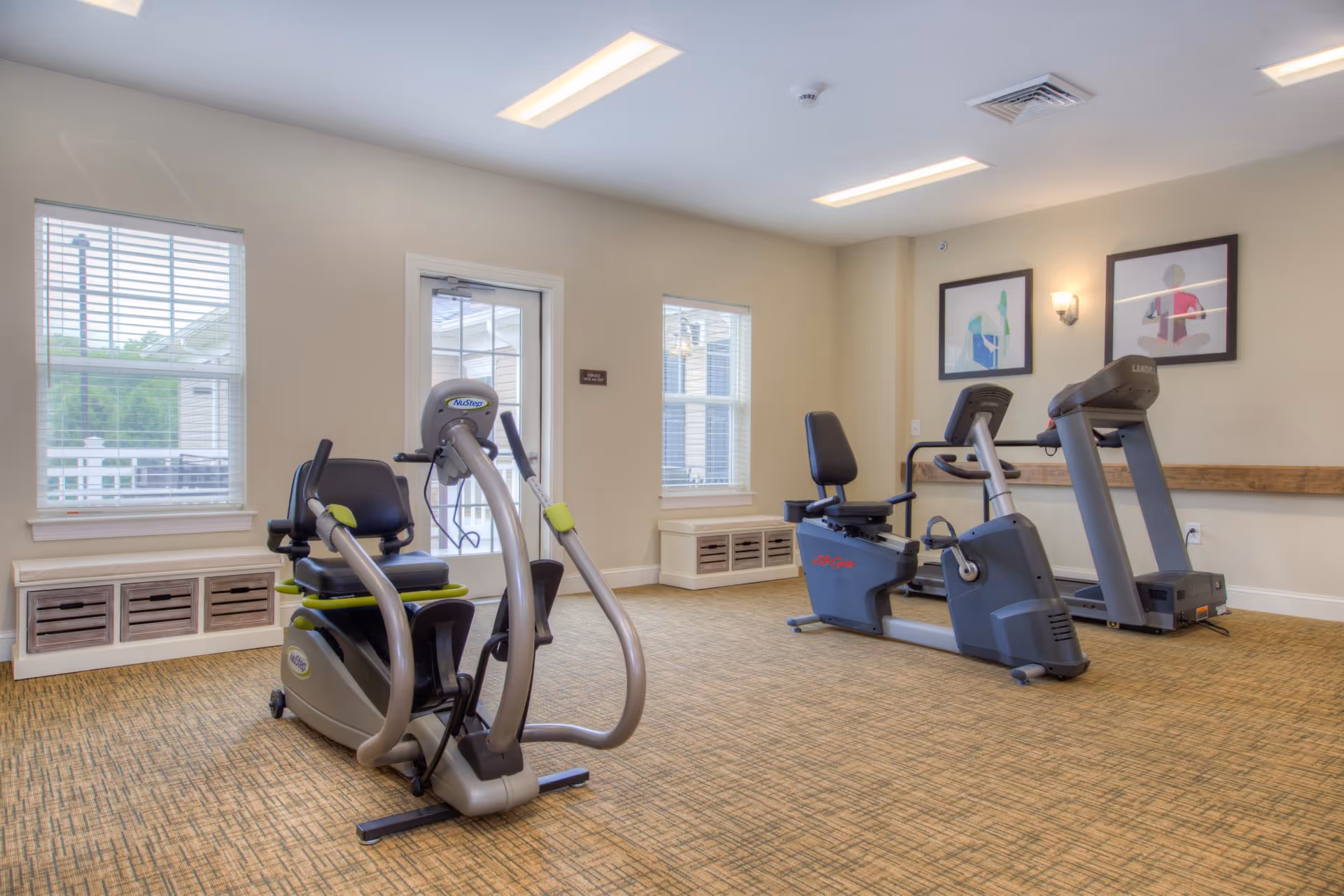 A bright exercise room with two recumbent stationary bikes and a treadmill. The room has beige walls, carpeted flooring, two windows with white blinds, and framed artwork on the wall. There is a door leading outside and ceiling lights providing illumination.