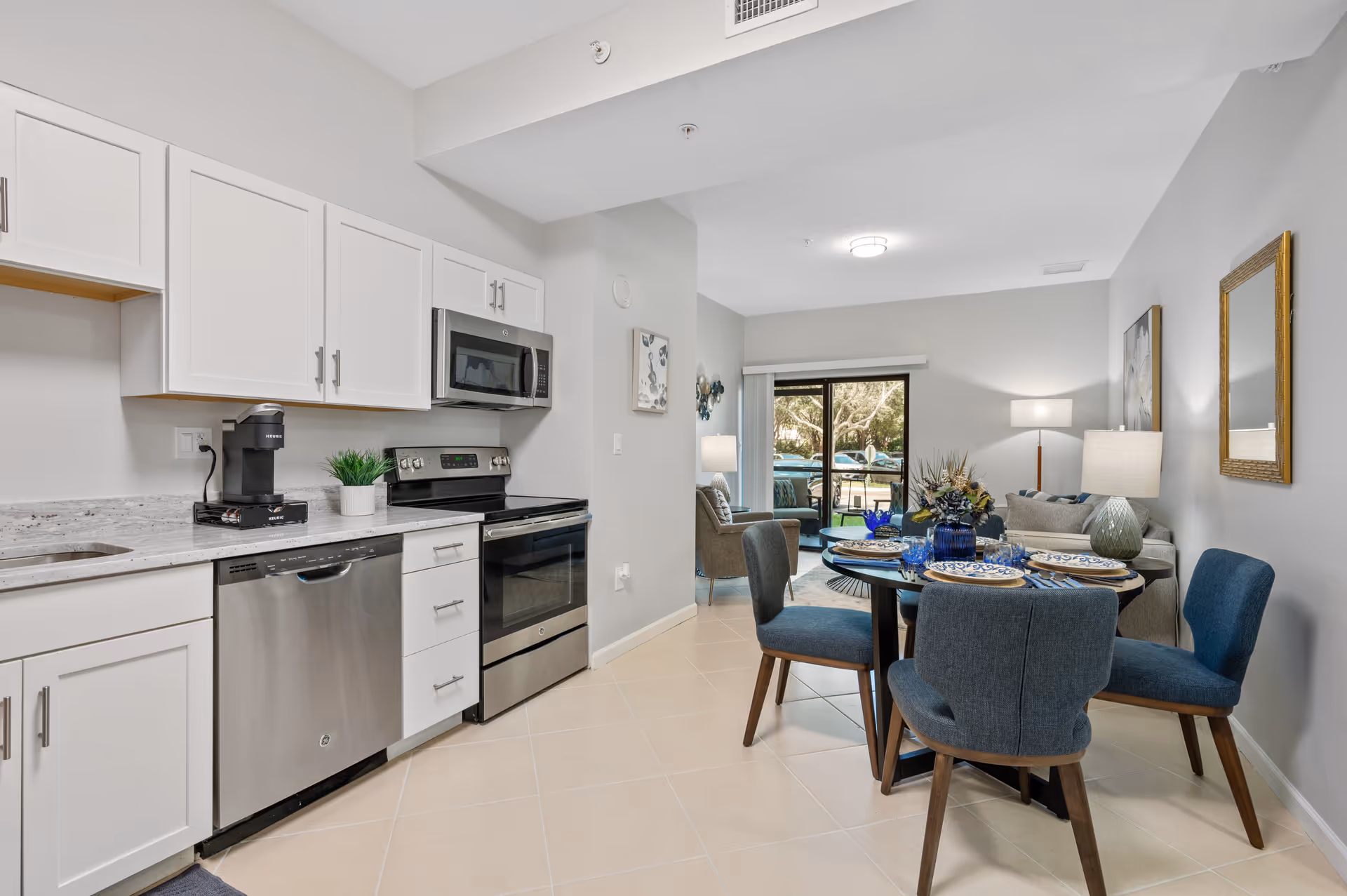 A modern open-concept kitchen and dining area in a senior living facility. The kitchen features white cabinets, a stainless steel dishwasher, oven, and microwave, with a coffee maker on the counter. The dining area has a round table set with blue and white plates, blue glasses, and blue upholstered chairs. In the background, there is a living room with a sofa, armchair, lamps, and a sliding glass door leading outside.
