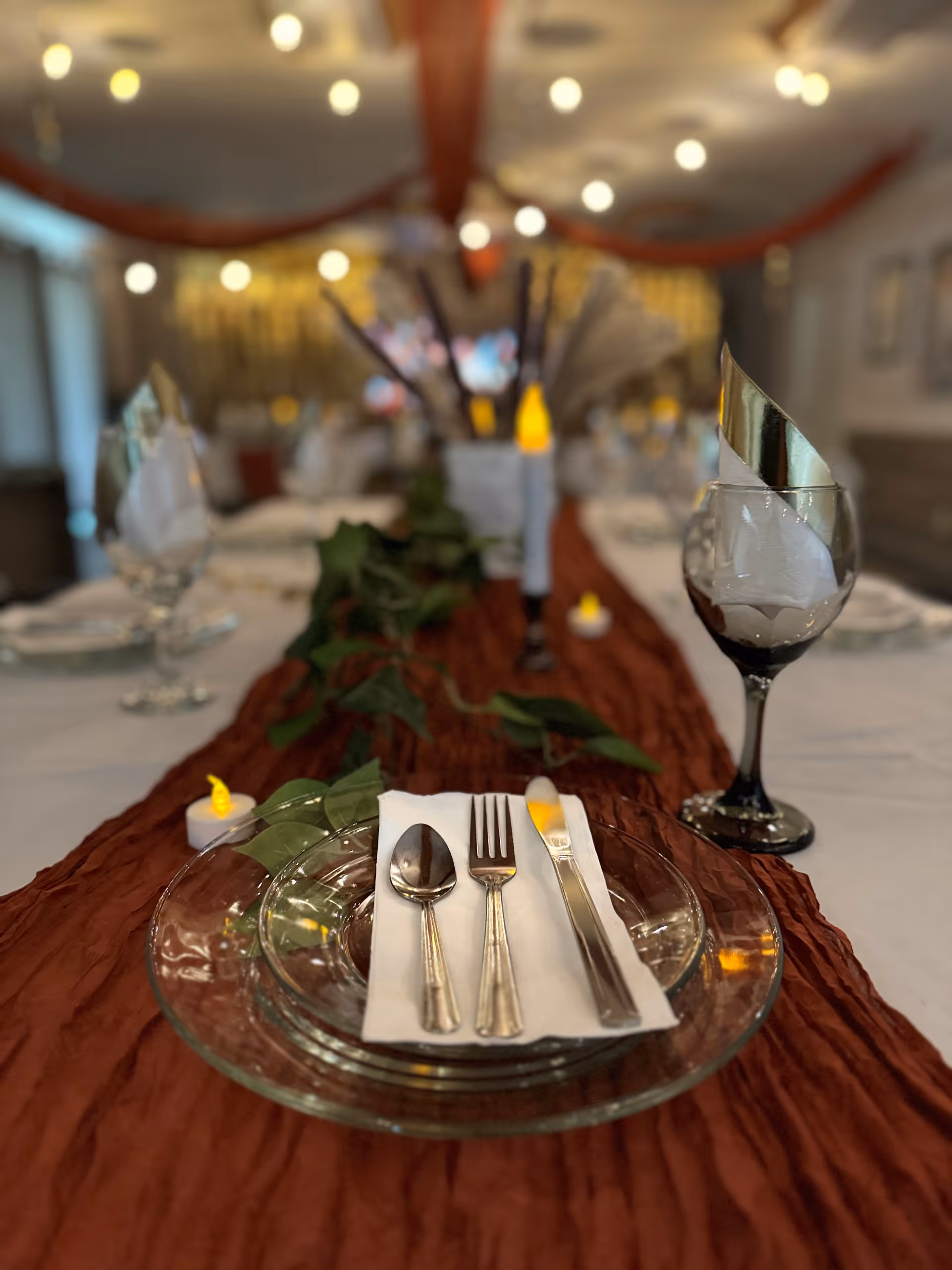 Close-up view of a dining table set with clear glass plates, silverware on a white napkin, a wine glass with a folded napkin inside, and a brown table runner decorated with green leaves and small LED candles in a softly lit room.