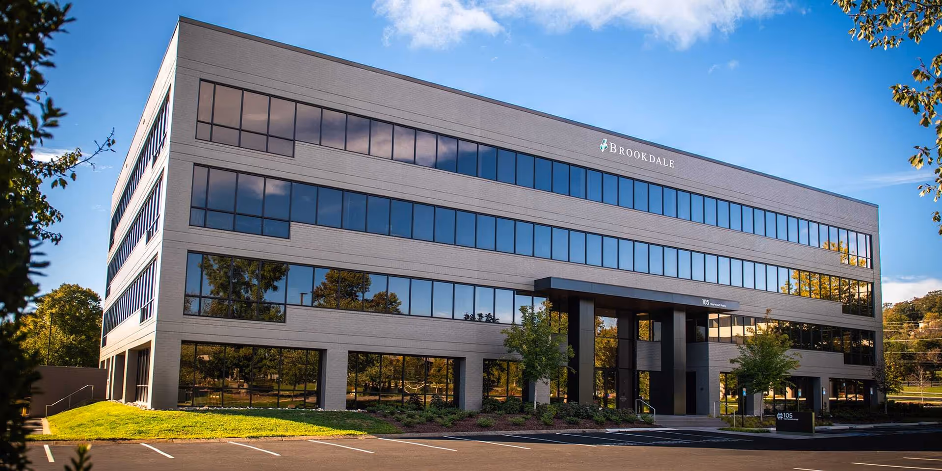 Exterior view of a modern four-story office building with large reflective windows and the Brookdale Senior Living Inc. logo on the top right corner. The building is surrounded by green grass, small trees, and a parking lot in front under a clear blue sky.