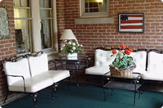 A cozy indoor seating area with white cushioned wrought iron chairs and a loveseat arranged around a glass-top coffee table. A potted plant with red flowers sits on the table. The walls are made of red brick, and there is a framed American flag hanging on the wall. A table lamp and another potted plant are on a side table next to the loveseat.