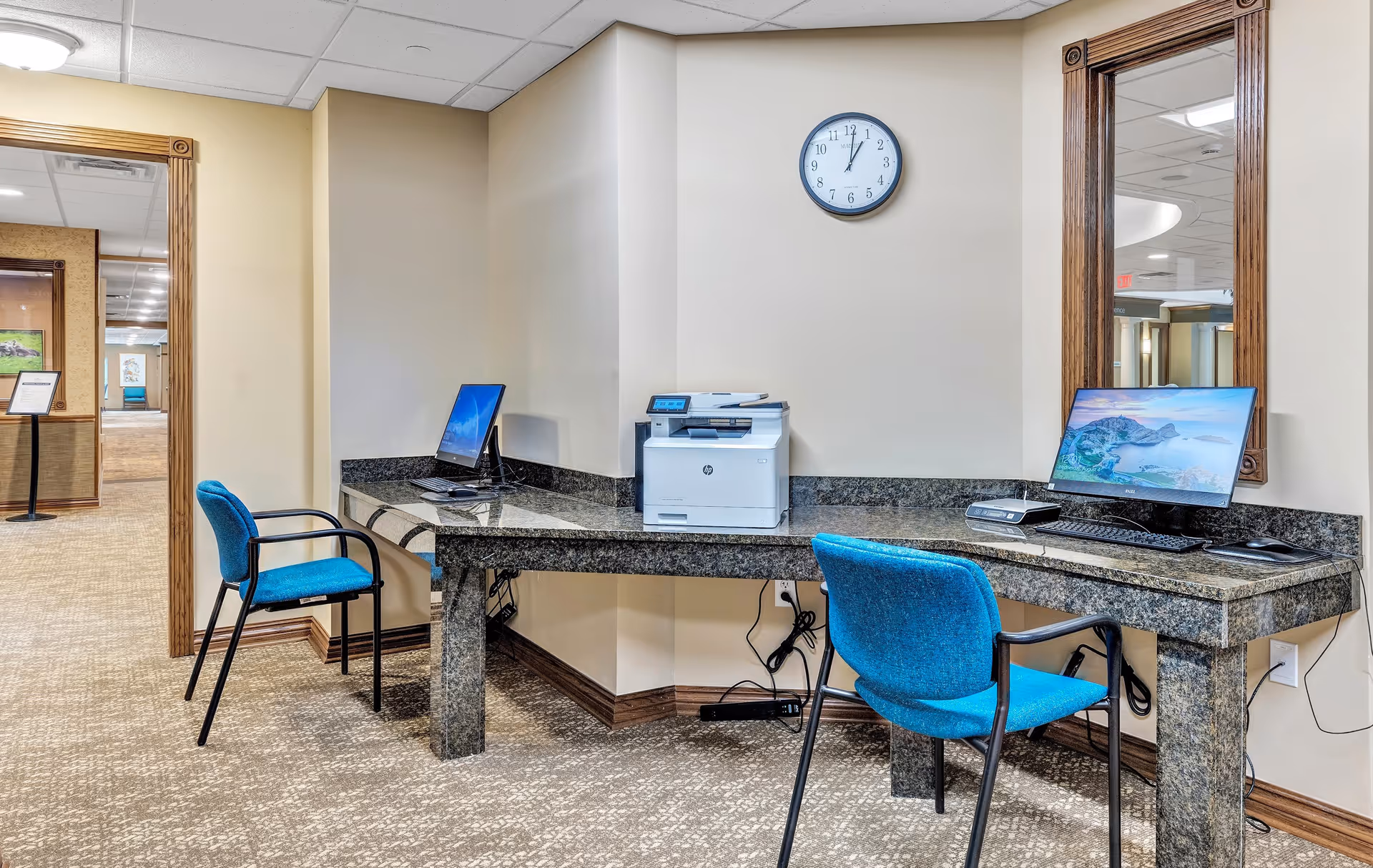 A small office corner in a retirement community with two blue chairs and a granite countertop desk. The desk holds two desktop computers and a printer. A wall clock shows the time as 12:05. There is a large mirror on the wall reflecting part of the hallway.