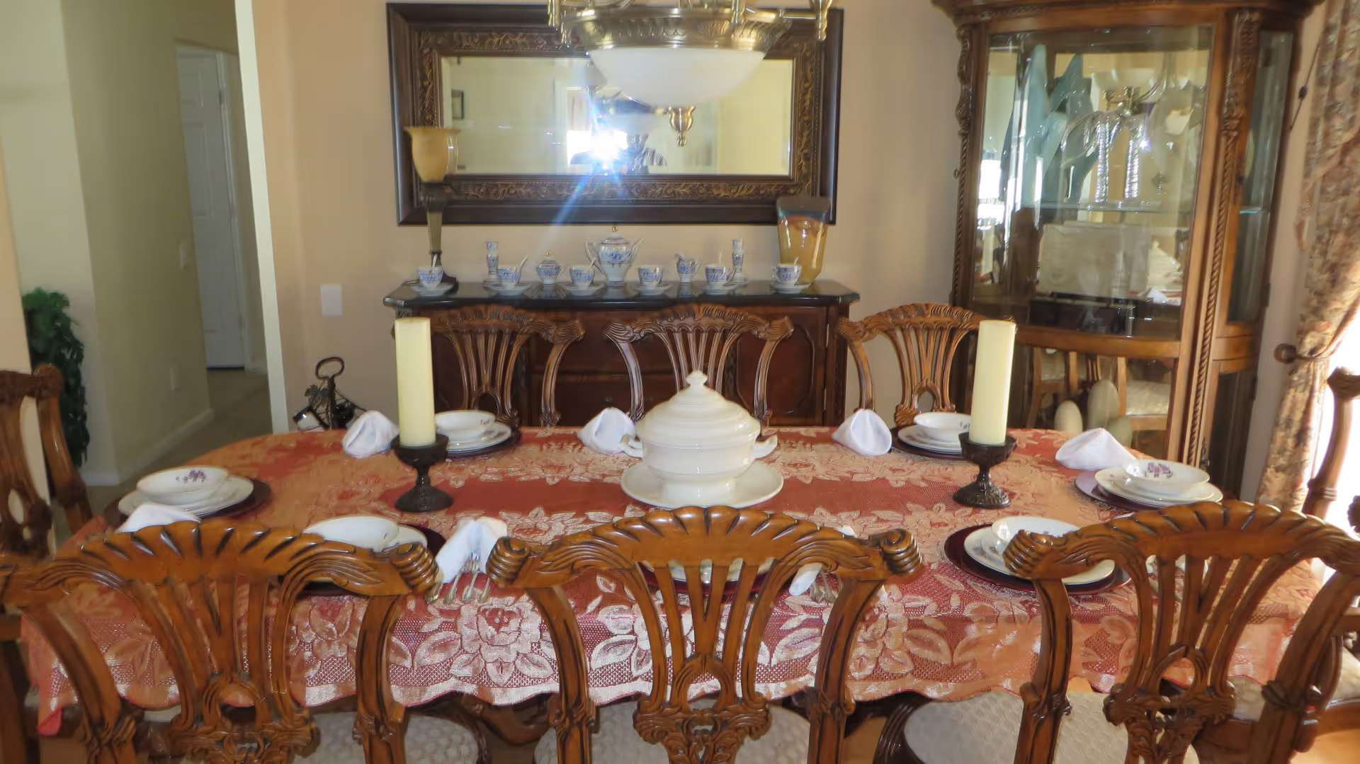 A formal dining room with a wooden dining table covered with a red floral tablecloth. The table is set with plates, napkins, and two large candles in holders. There is a white tureen in the center of the table. Behind the table is a wooden sideboard with a tea set and a large ornate mirror above it. To the right is a glass display cabinet with decorative items inside. The room has beige walls and a window with floral curtains.