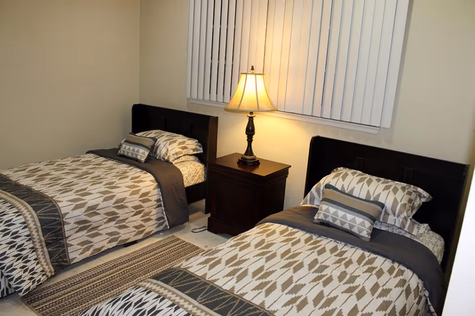 A bedroom with two twin beds featuring matching patterned bedding in neutral tones. Between the beds is a dark wooden nightstand with two drawers, topped by a lit table lamp. A window with vertical blinds is behind the nightstand, and a patterned rug lies on the floor in front of the beds.
