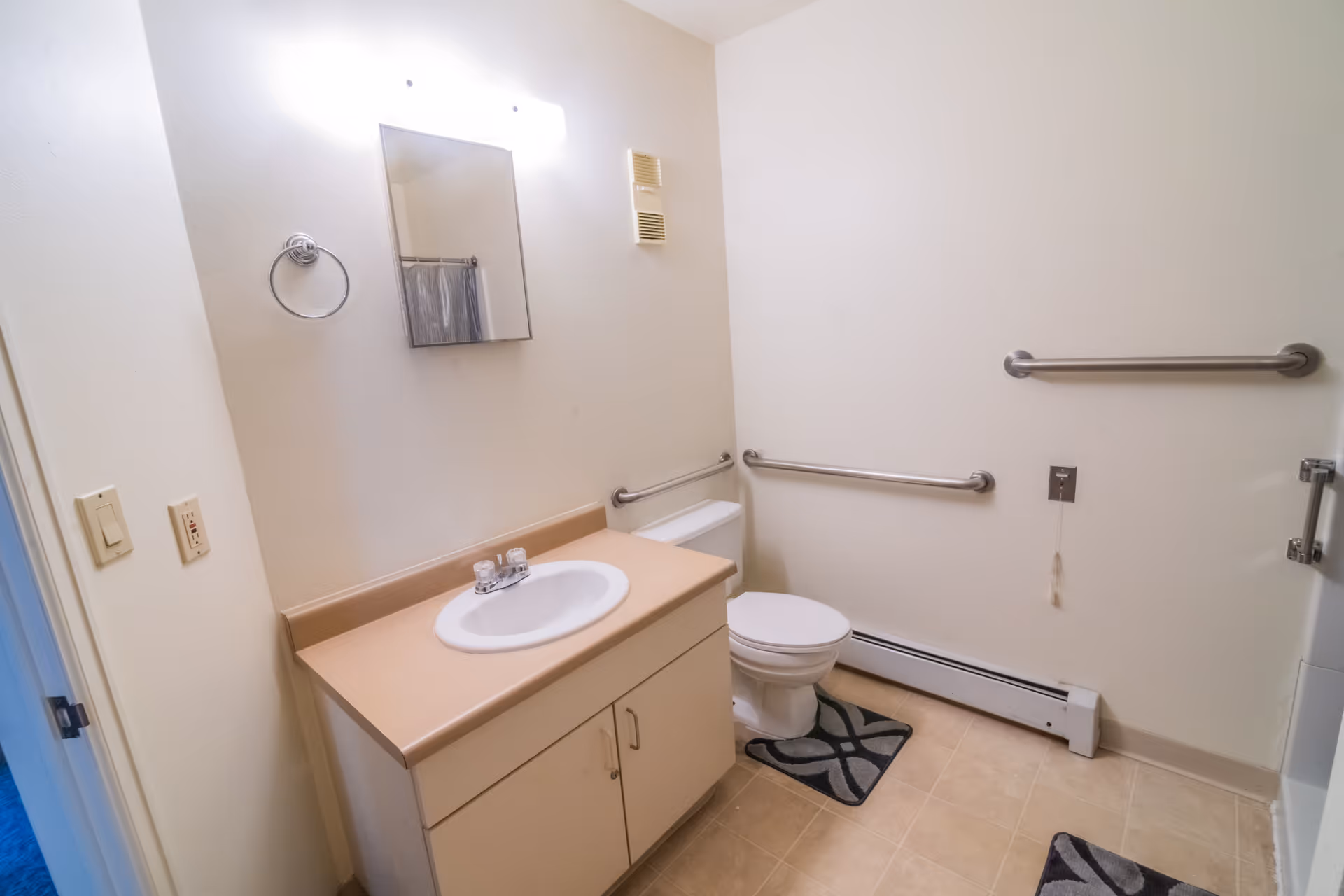 A clean bathroom with beige countertop and white sink, a mirror above the sink, a toilet with grab bars on the walls around it, and beige tiled floor with two patterned rugs.
