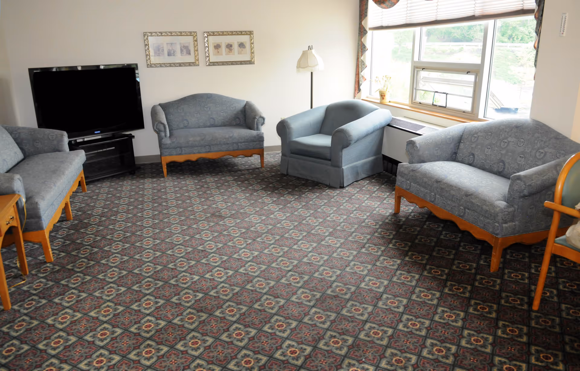 A living room with patterned carpet, three blue upholstered sofas, a blue armchair, a flat-screen TV on a stand, a floor lamp, two framed pictures on the wall, and a window with blinds partially open letting in natural light.