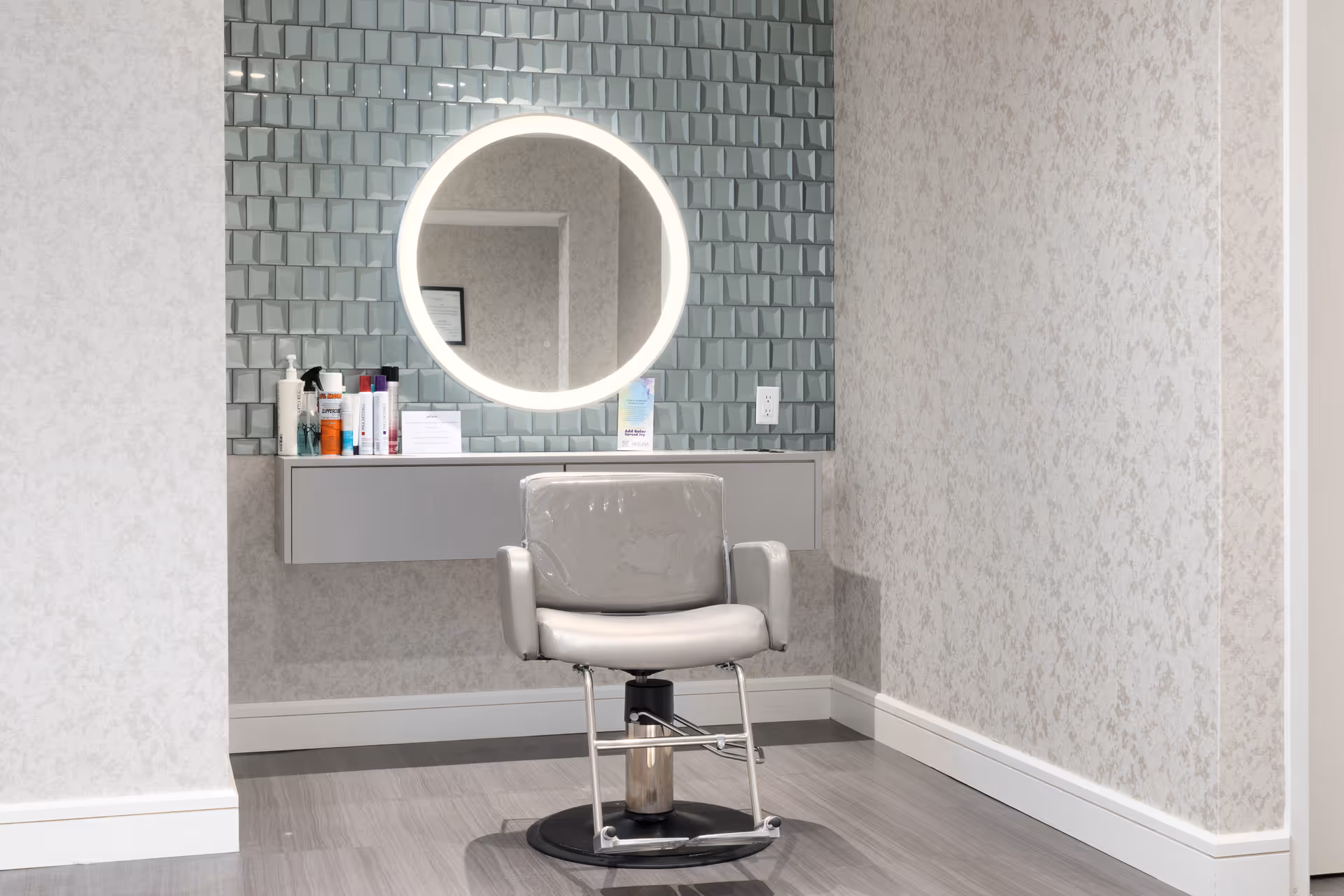 A modern salon or grooming area with a single gray salon chair in front of a wall-mounted floating shelf holding various hair and beauty products. Above the shelf is a round mirror with built-in lighting, set against a textured blue-gray tiled wall. The surrounding walls have a subtle floral pattern wallpaper and the floor is a light gray wood or laminate.