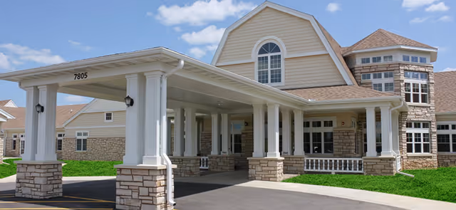 Front exterior of the Pride TLC Therapy & Living Campus showing a covered entrance with white columns, stone facade and manicured lawn under a blue sky.