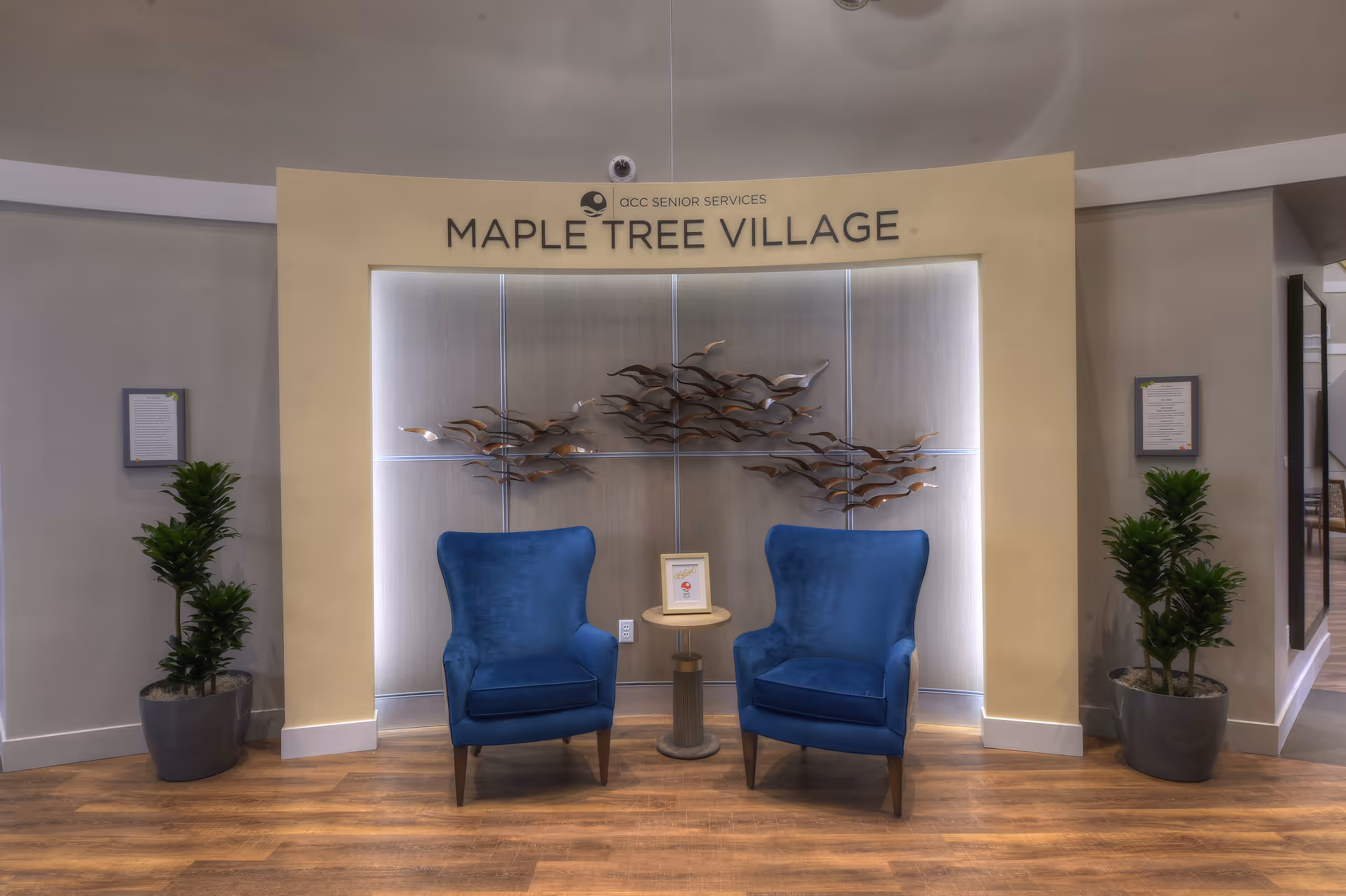 A cozy seating area in a senior living community featuring two blue armchairs with a small round table between them, placed against a wall with a decorative metal art installation of flying birds. The wall has a sign above reading 'ACC Senior Services Maple Tree Village'. Two potted plants flank the seating area on either side.