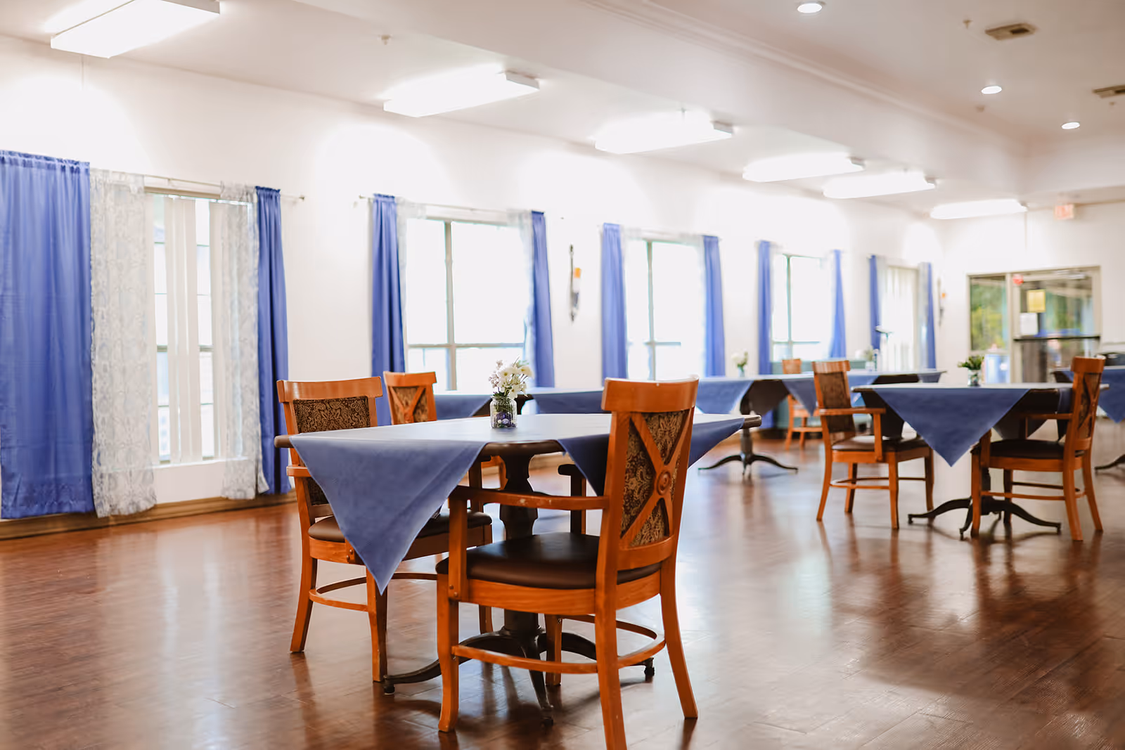 A bright dining room with wooden floors and several wooden tables covered with blue tablecloths. Each table has wooden chairs with patterned backs and small flower arrangements in the center. Large windows with blue and white curtains line the walls, allowing natural light to fill the room.