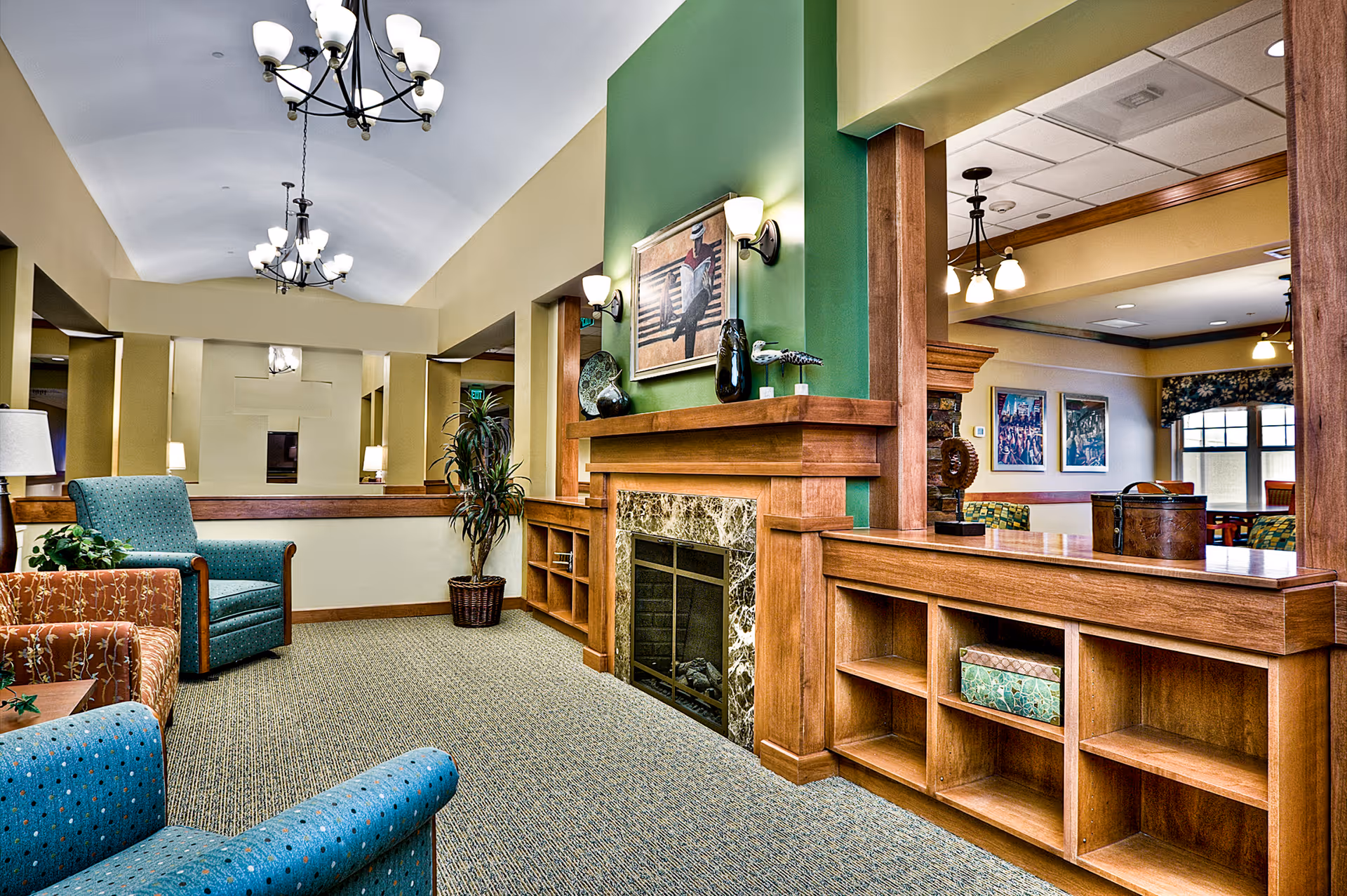 A cozy living room area in a senior living facility with comfortable upholstered chairs in blue and orange patterns, a green accent wall with a fireplace surrounded by wooden shelves, decorative vases, and a painting above the fireplace. The room has carpeted flooring, warm lighting from chandeliers and wall sconces, and an open view into an adjacent room with framed artwork on the walls.