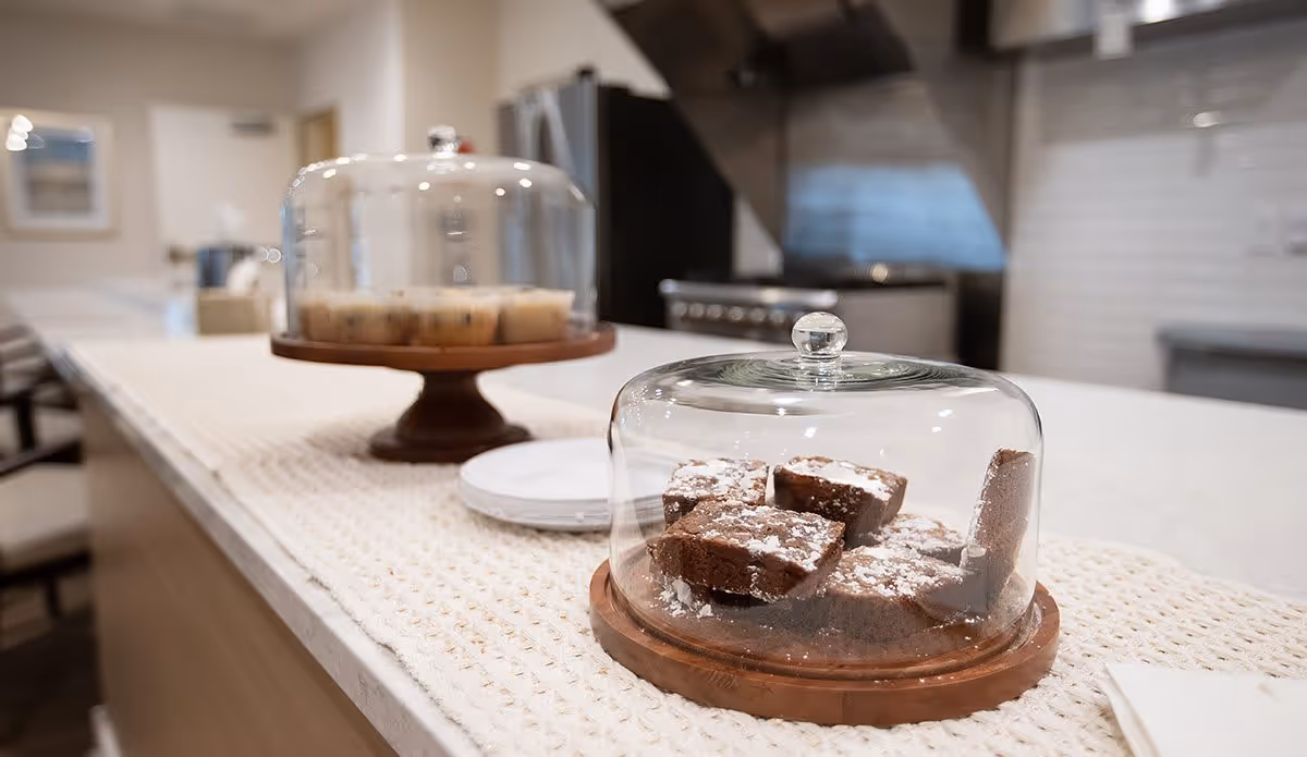 Close-up view of a kitchen counter with two wooden cake stands covered by glass domes. One stand holds several brownies dusted with powdered sugar, and the other holds muffins. The background shows a modern kitchen with a stove and stainless steel appliances.