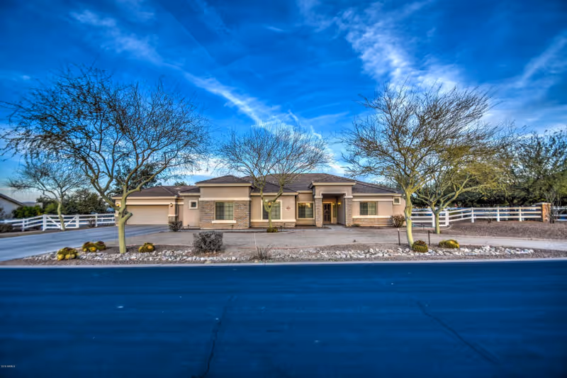 Single-story residential building with a beige exterior and a tiled roof, surrounded by leafless trees and desert landscaping. The building has a driveway and a white fence in the background under a blue sky with scattered clouds.