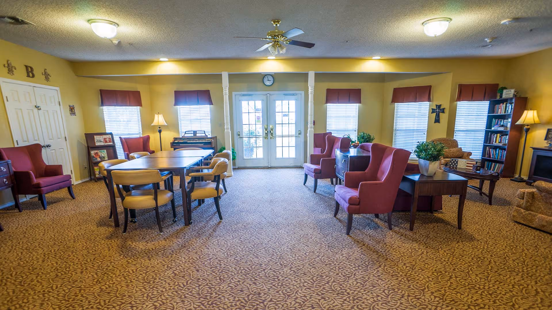 A cozy common area in a senior living facility with yellow walls and patterned carpet. The room features several red upholstered armchairs, a wooden table with chairs, a bookshelf filled with books, a floor lamp, and a small electric fireplace. There are multiple windows with red valances and a set of double glass doors at the far end of the room. The ceiling has a fan and light fixtures.