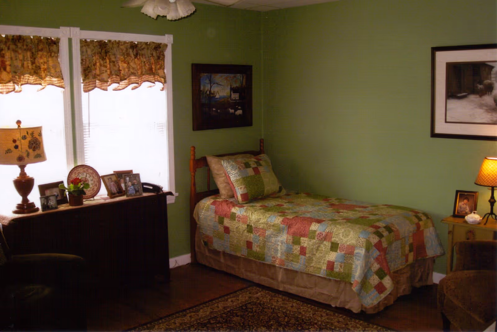 A cozy bedroom with green walls featuring a single bed with a colorful patchwork quilt and matching pillow. There is a wooden nightstand with a lamp and framed photo, a dark wooden dresser with a lamp, framed photos, a decorative plate, and a small potted plant. Two windows with floral valances allow light into the room. The floor has a patterned rug and there are framed pictures on the walls.