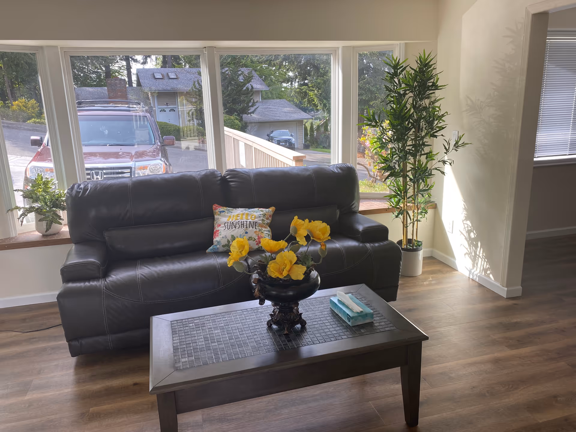 A living room with a black leather sofa that has a colorful pillow reading 'Hello Sunshine'. In front of the sofa is a wooden coffee table with a decorative vase holding yellow flowers and a box of tissues. Large windows behind the sofa show a view of parked cars and houses outside. There is a tall green plant in a white pot to the right of the sofa, and the room has wooden flooring and light-colored walls.
