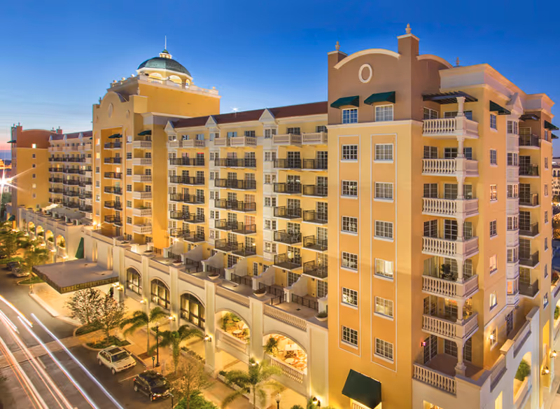 Exterior view of a large multi-story residential building at dusk with balconies, arched windows, and a dome on the roof. The building is illuminated with warm lights and palm trees line the street below.
