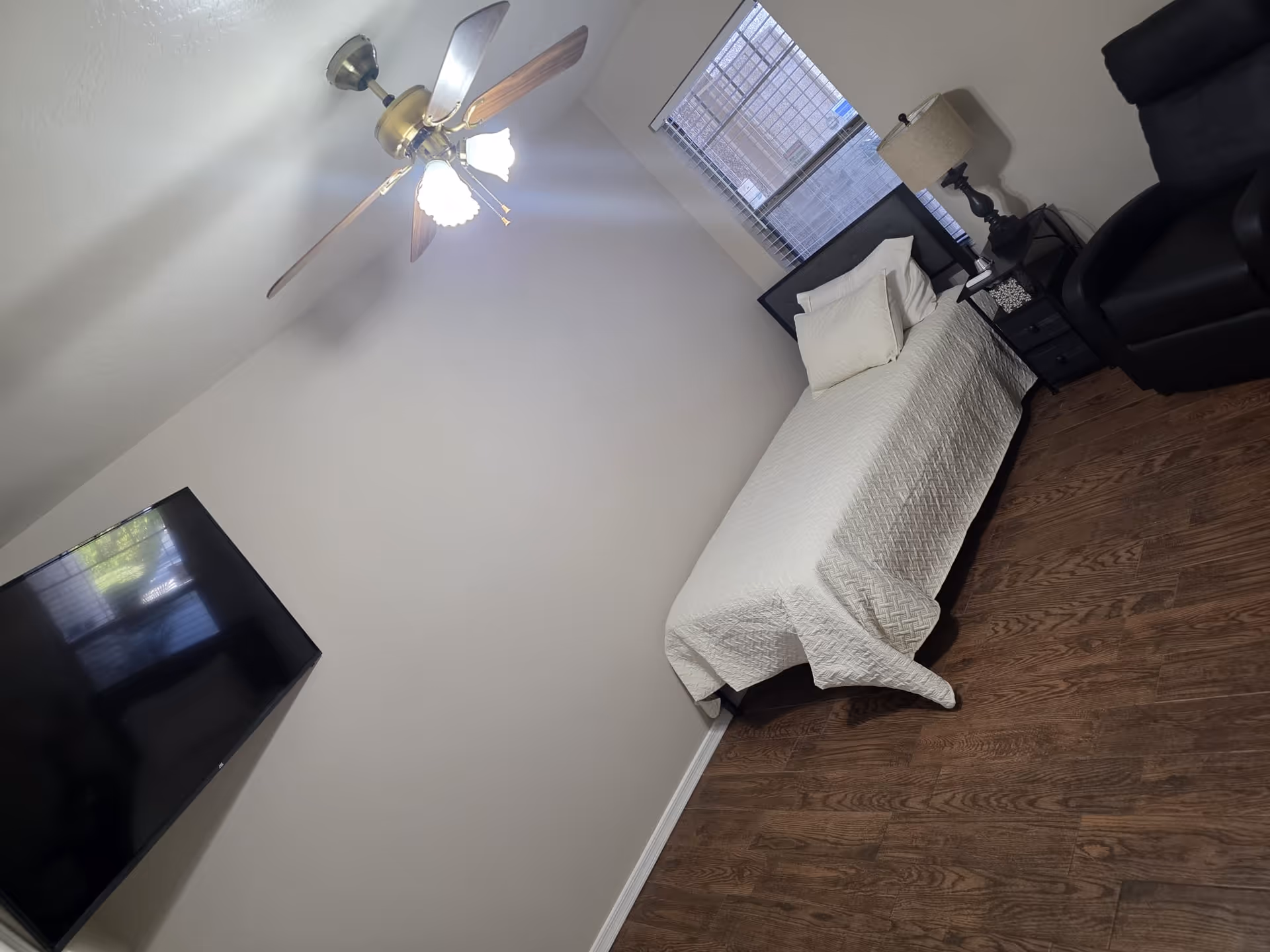 Small furnished bedroom with a single bed, wall-mounted TV, nightstand with lamp, and armchair on wood flooring.