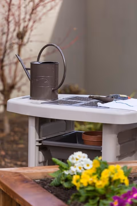 A gardening table with a brown watering can, a small hand trowel, and a white cloth on top. In the foreground, there is a raised garden bed with blooming yellow, white, and purple flowers. The background shows a blurred outdoor setting with a tree and a beige wall.
