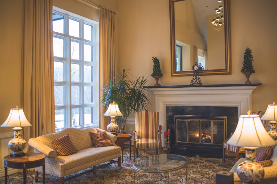 A cozy living room with a beige sofa, striped armchairs, a glass coffee table, and two side tables with decorative lamps. There is a large window with beige curtains letting in natural light, a fireplace with a mirror above it, and some decorative plants and candles around the room.