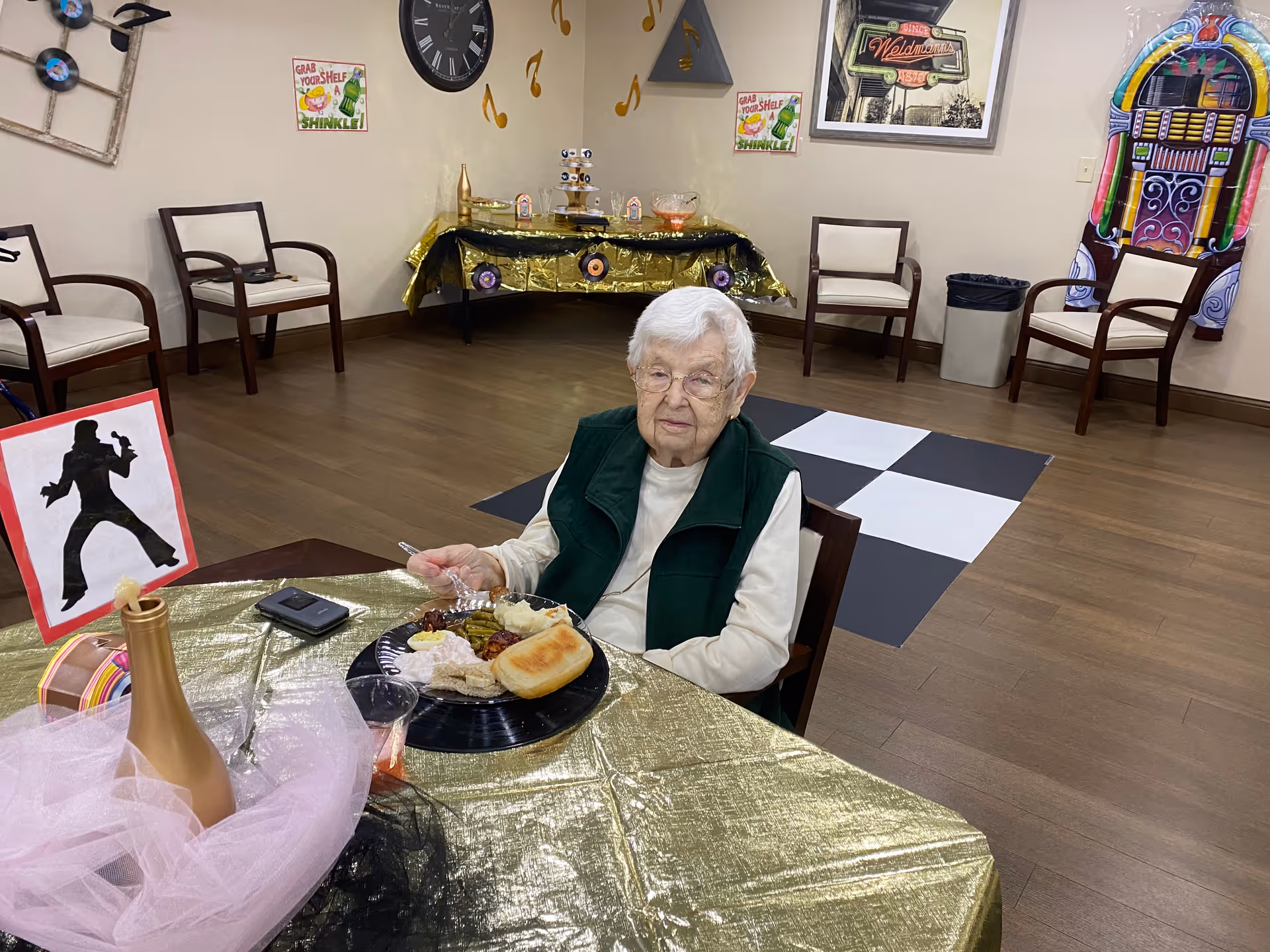 An elderly woman sits at a decorated table with a plate of food in a communal activity/dining room.