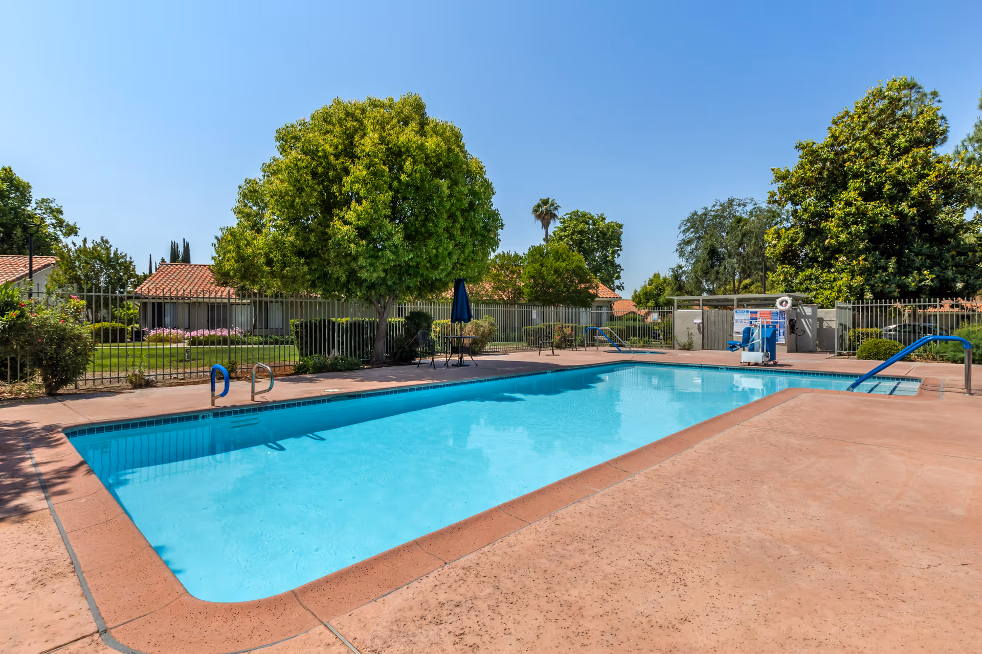 Outdoor swimming pool with clear blue water surrounded by a concrete deck. There are trees, bushes, and a metal fence enclosing the pool area. In the background, there are single-story buildings with red tile roofs under a clear blue sky.