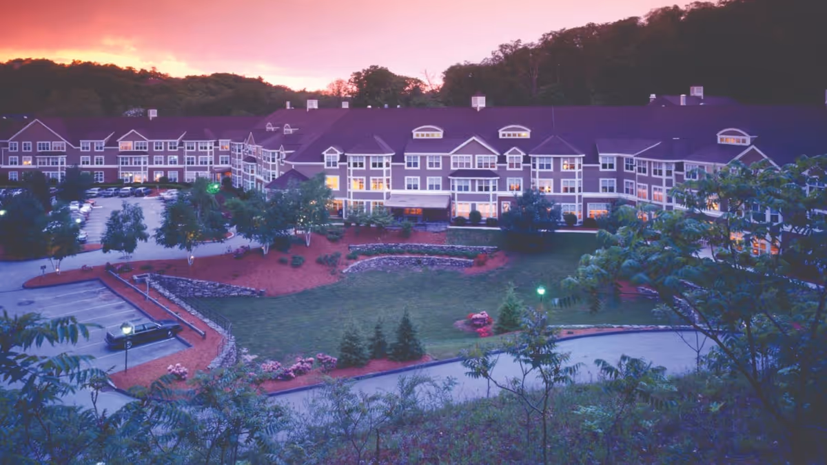 Exterior view of The Gables at Winchester senior living facility at sunset, showing a large multi-story building with many windows illuminated, surrounded by landscaped gardens, trees, and a parking area.