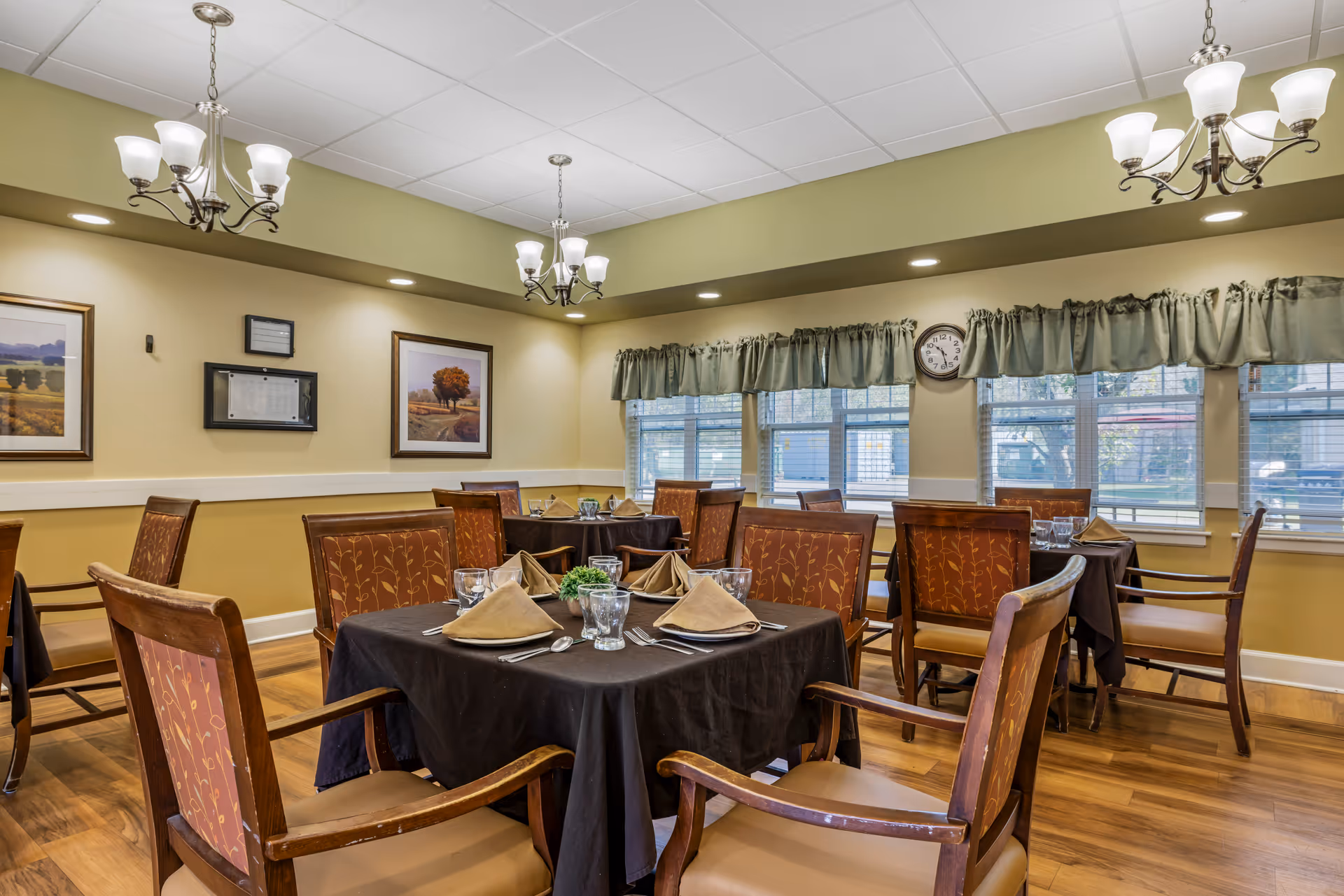 A dining room with several tables covered in dark tablecloths, each set with folded beige napkins, glasses, and silverware. The room has wooden chairs with patterned upholstery, hardwood floors, green walls, framed landscape paintings, and windows with green valances. Two chandeliers with multiple lights hang from the ceiling.