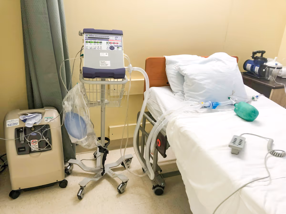 A hospital or healthcare facility room with a bed equipped with white sheets and pillows. Medical equipment including a ventilator and oxygen supply machine are positioned next to the bed. Tubes and medical devices are connected to the bed, indicating a patient care setting.