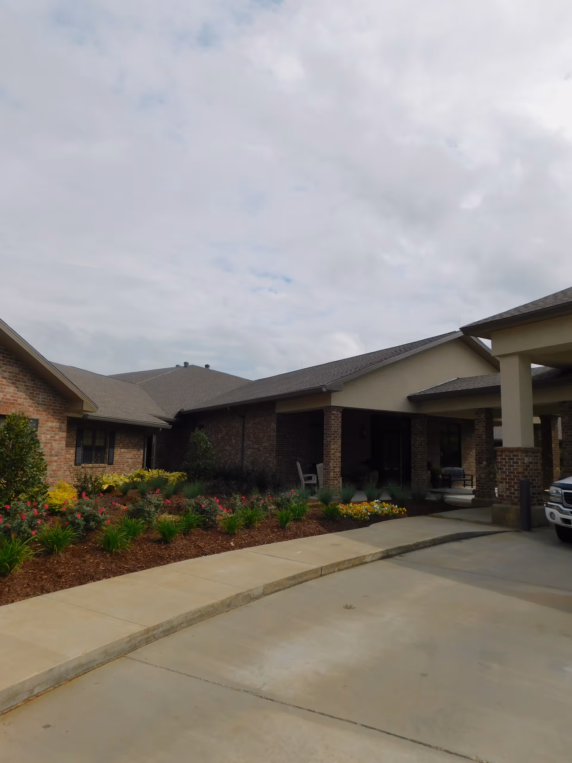 Exterior view of Landmark Nursing Center Hammond showing a brick building with a covered entrance, landscaped flower beds with various plants and flowers, and a concrete driveway under a cloudy sky.
