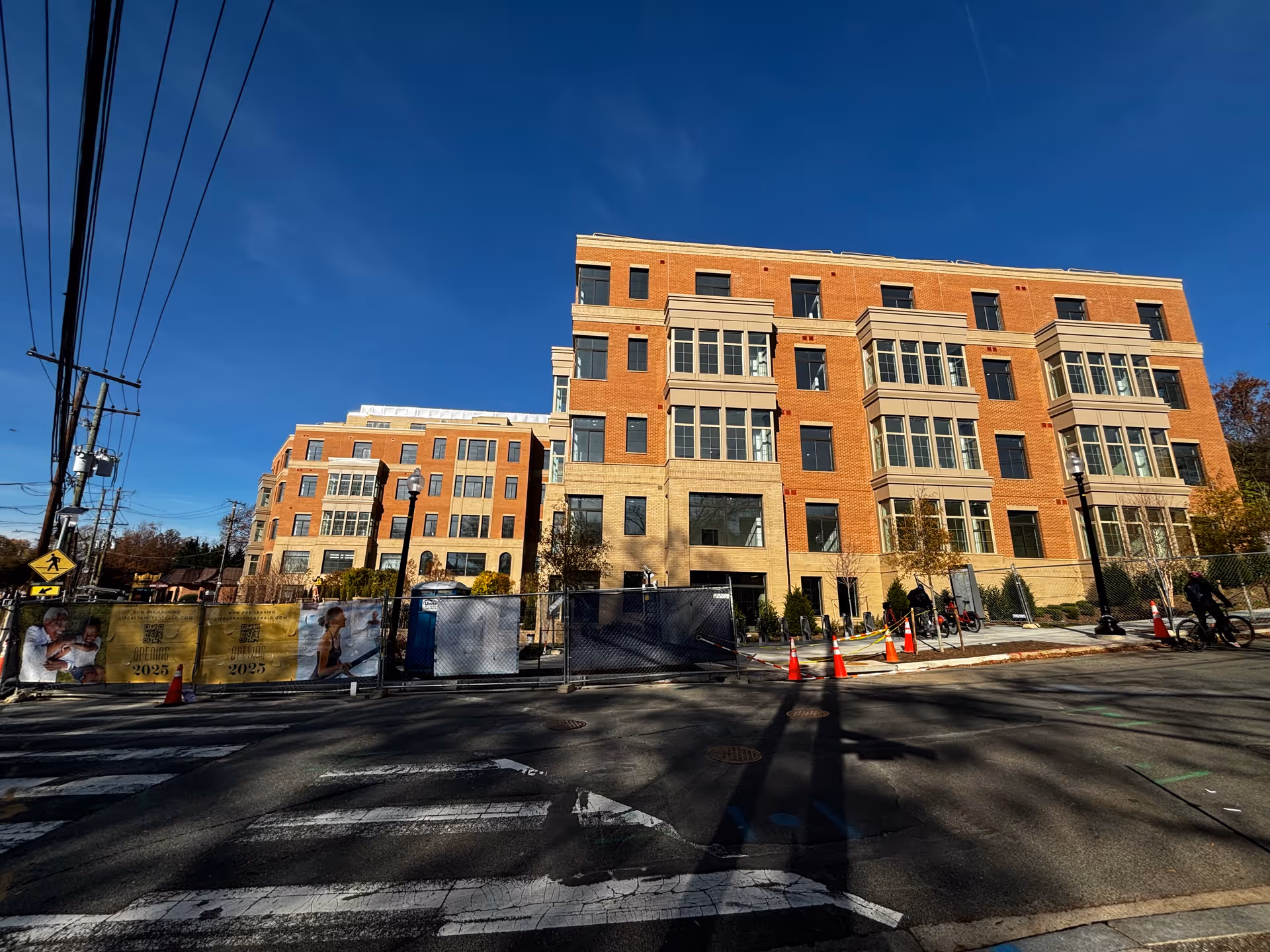 Exterior view of a multi-story brick building under construction with large windows and beige accents, surrounded by a construction fence and orange traffic cones, under a clear blue sky.
