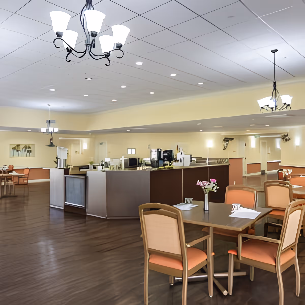 Spacious dining area with multiple tables and chairs, soft yellow walls, and modern chandeliers hanging from a white ceiling. A counter area with coffee machines and other equipment is visible in the background. The floor is dark wood, and the tables are set with napkins and small flower vases.